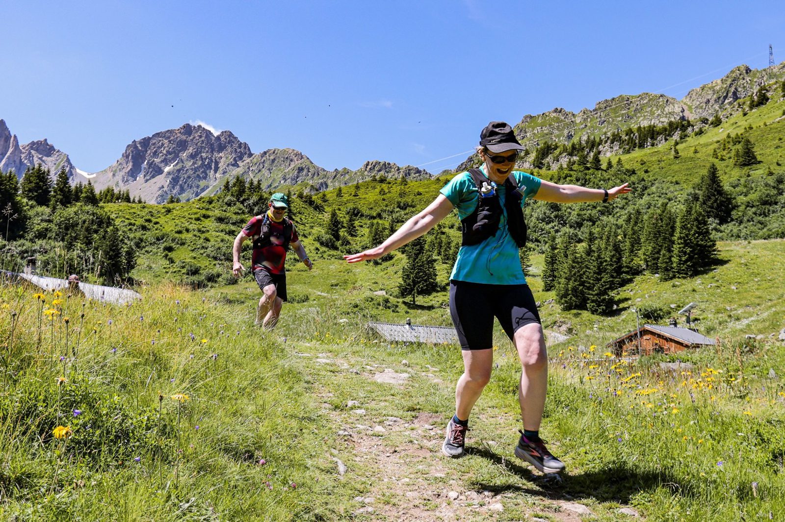 Two hikers, one man and one woman, descend a narrow mountain trail surrounded by green hills and wildflowers. Both wear athletic clothing and caps. The woman leads with arms out for balance, while the man follows close behind. Snow-capped peaks and a clear blue sky are in the background.
