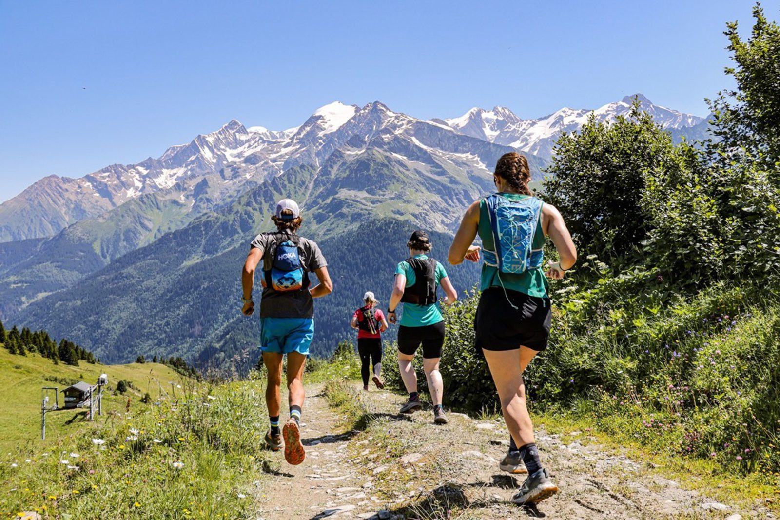 Four people run on a trail through a mountainous landscape on a sunny day. They wear running gear and carry hydration packs. Snow-capped peaks and lush green valleys are in the background, and the sky is clear blue. Wildflowers dot the grassy terrain.