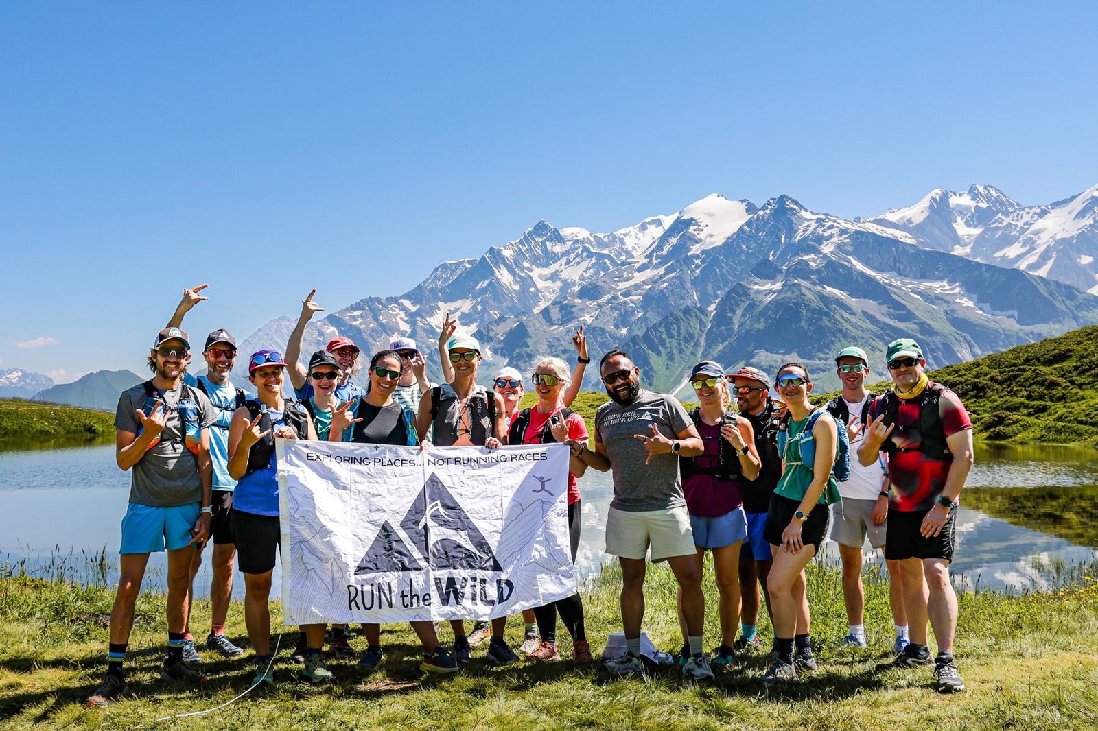 A group of people dressed in athletic wear standing outdoors, posing with a banner that reads "Run the Wild." Snow-capped mountains and a lake are visible in the background under a clear blue sky. The group appears to be celebrating or commemorating an event.