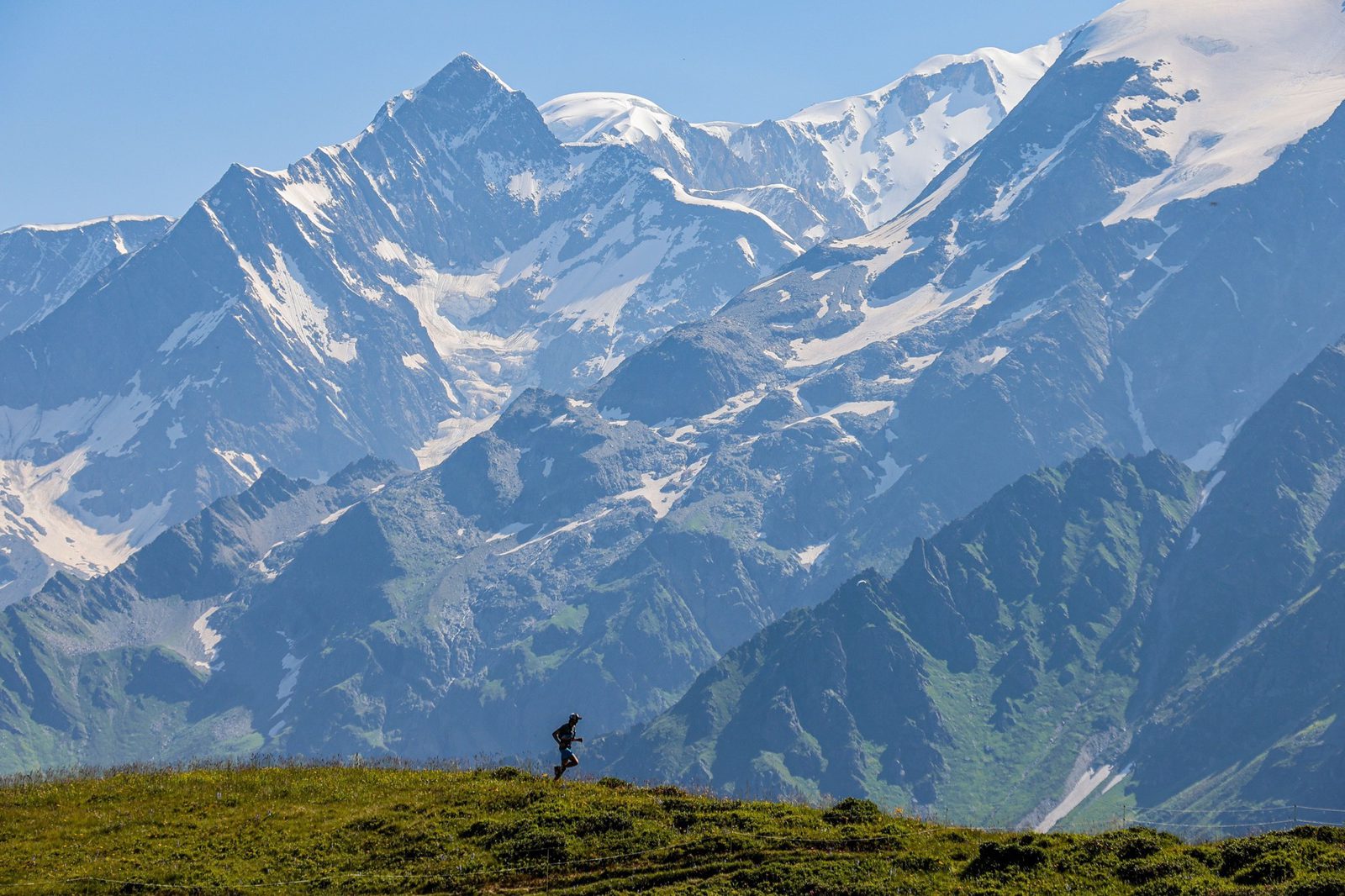 A lone mountain biker rides along a grassy ridge with a breathtaking view of towering, snow-capped peaks in the background. The sky is clear, highlighting the majestic mountain scenery.