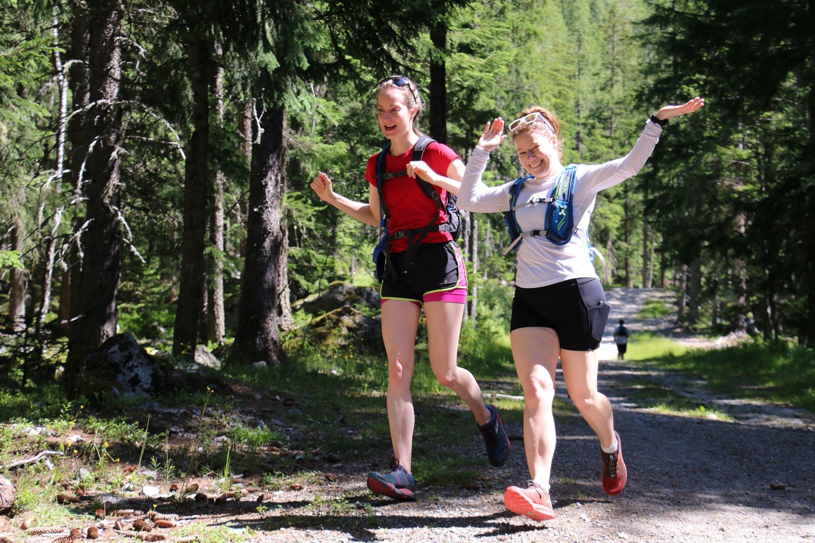 Two people are jogging on a forest trail, smiling and lifting their arms in joy. They are wearing athletic gear, including shorts and tops, and are surrounded by tall trees and dappled sunlight filtering through the foliage.