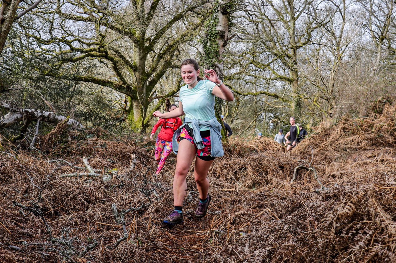 A woman in athletic wear, carrying a red backpack, runs on a wooded trail. She is surrounded by dry, brown foliage and leafless trees. Other runners can be seen in the background. The scene is bright and appears to be during autumn or early spring.