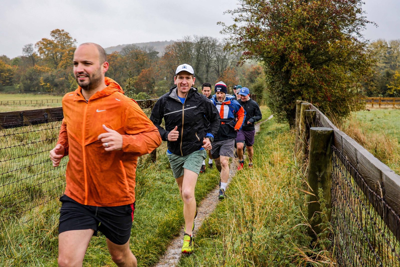A group of people are running on a narrow dirt path along a fence in a countryside setting, with greenery and autumn-colored trees in the background. The lead runner wears an orange jacket, and others follow closely behind, smiling despite the overcast weather.
