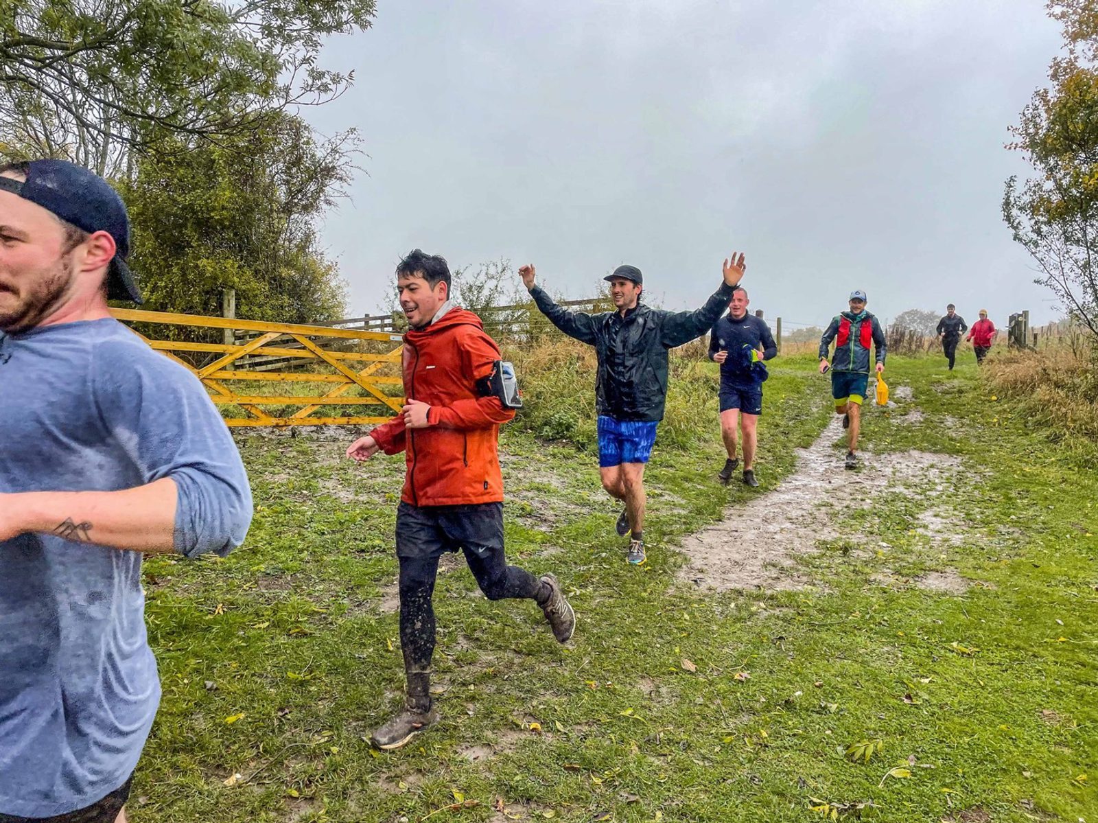 A group of people running on a muddy path in an outdoor setting. The runner in the foreground is in focus, wearing an orange jacket, black pants, and carrying a hydration pack. Other runners are seen in the background dressed in various outdoor attire.