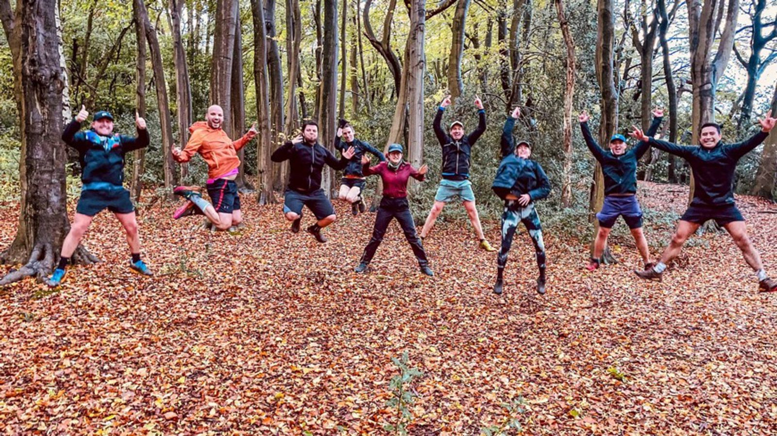 A group of people wearing athletic clothing are jumping in unison in a forest with autumn leaves covering the ground. They appear joyful, with arms and legs extended. Tall trees with yellowish-green leaves surround them, creating a vibrant, outdoor setting.