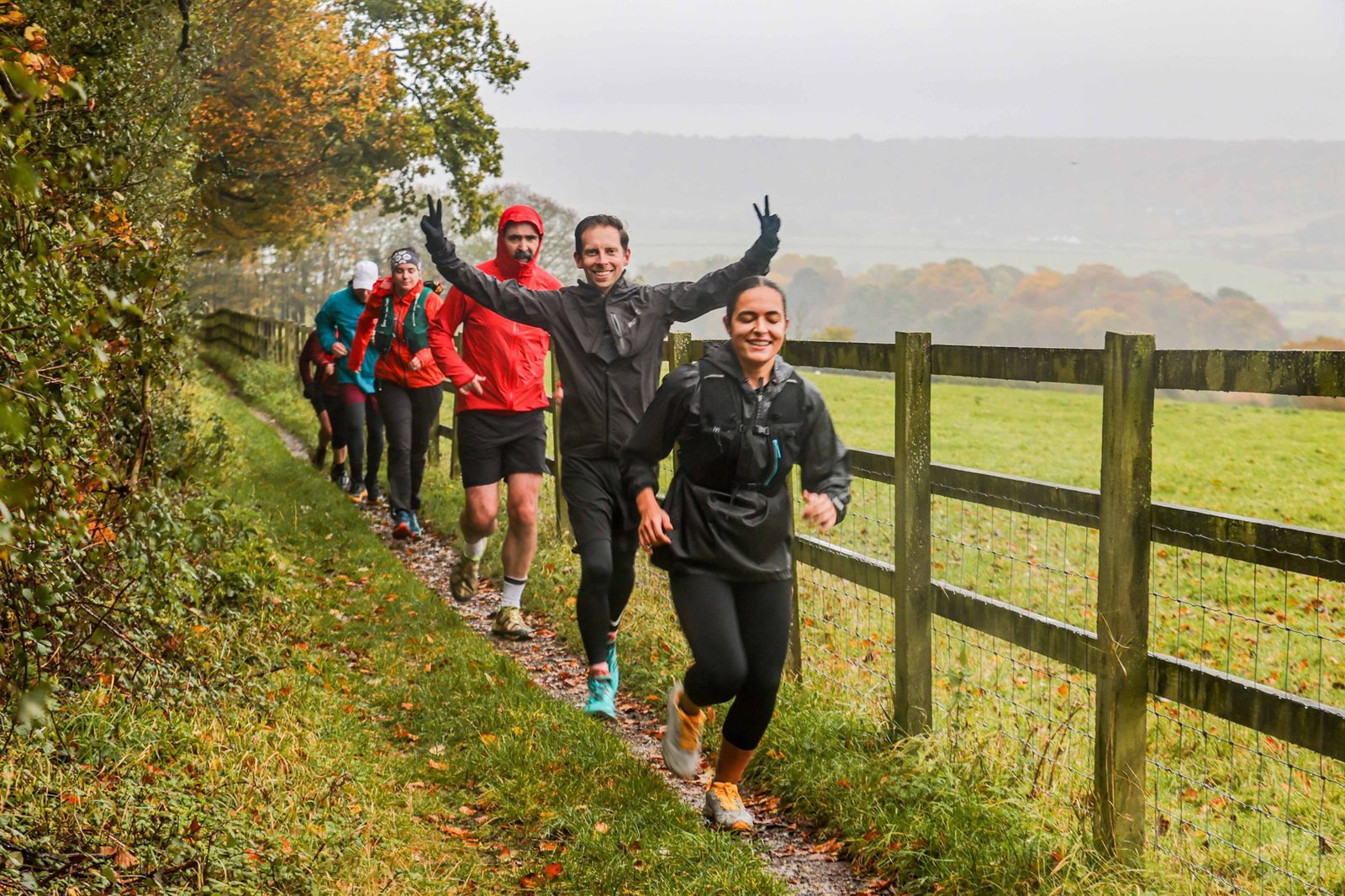 A group of people is jogging on a narrow path beside a wooden fence in a rural area. The lead jogger, a woman in a black outfit, smiles at the camera, while the man behind her raises his hands in a celebratory pose. The scene is autumnal with colorful foliage.