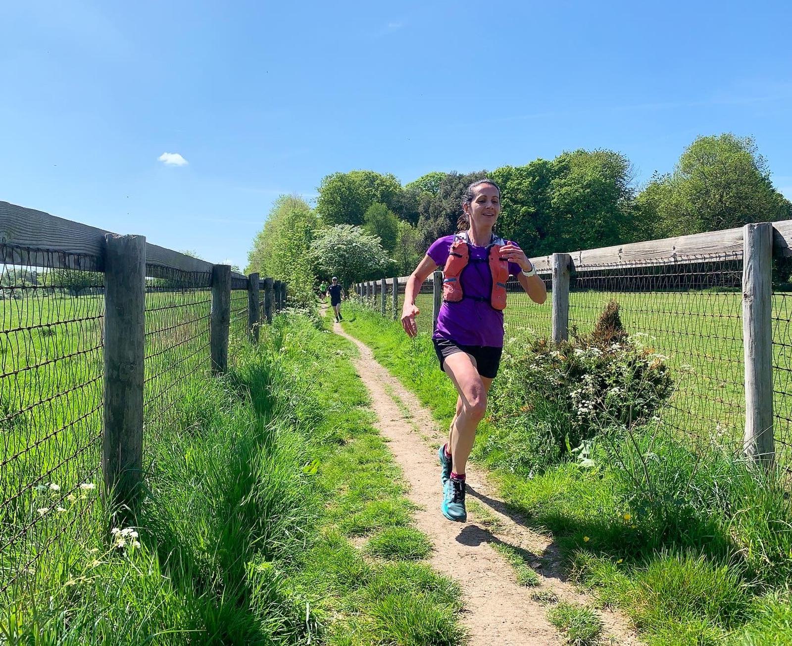 A person is running along a dirt path surrounded by greenery and wooden fences on a sunny day. They are wearing a bright purple outfit and carrying an orange hydration pack, smiling as they run. Another runner can be seen in the distance behind them.