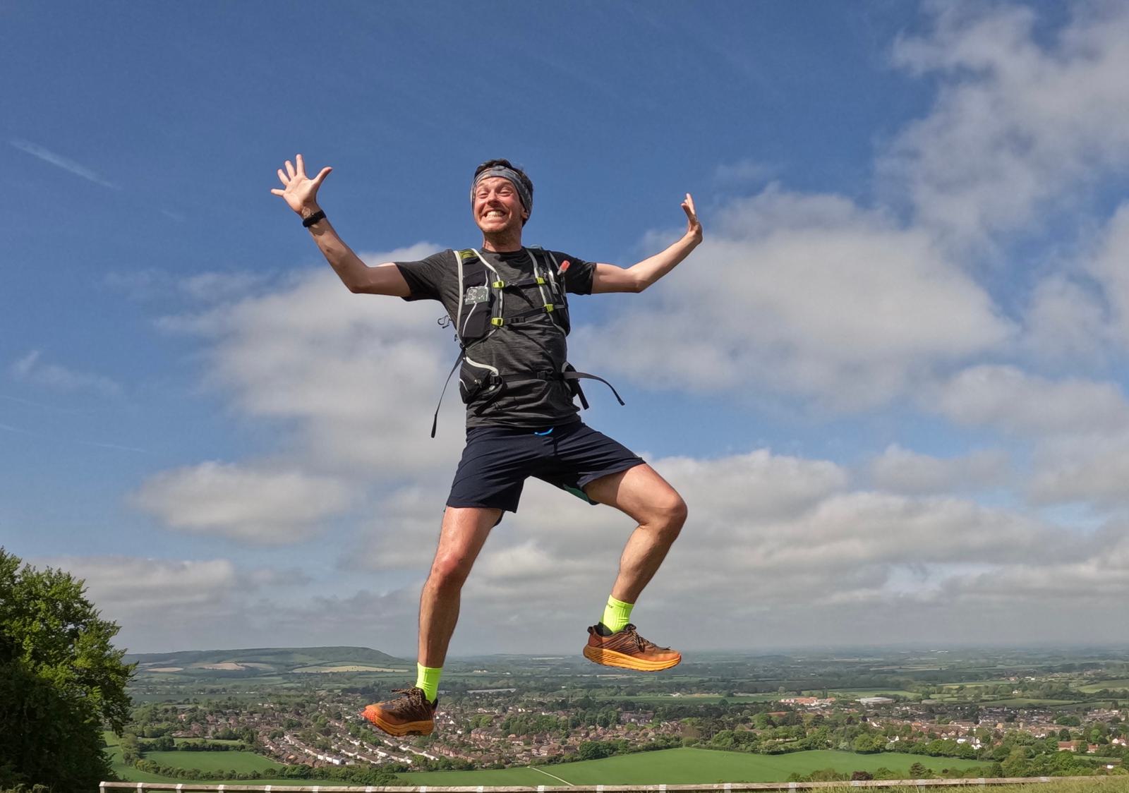 A man is leaping joyfully in the air on a sunny day, wearing a hydration backpack, black shorts, and bright running shoes. A scenic landscape with green fields, small towns, and a partly cloudy sky stretches out behind him.