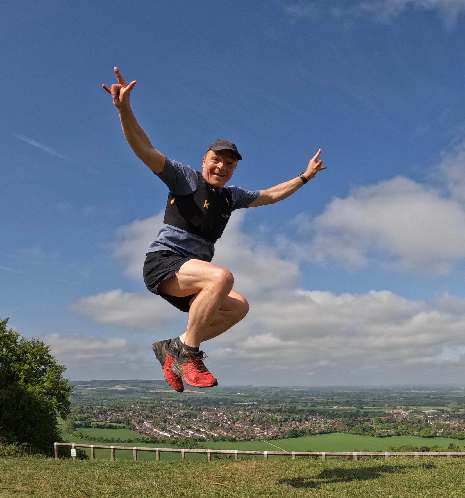 A man wearing a cap and athletic clothing, including red shoes and a black waist pack, jumps enthusiastically in the air against a scenic backdrop of a sprawling town and lush green fields under a blue sky with scattered clouds.