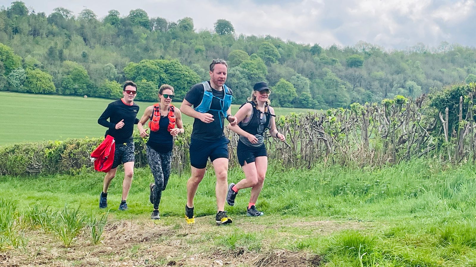 A group of four people wearing athletic gear run together through a lush green field with a wooded hillside in the background. The sky is partly cloudy. The runners look focused and are positioned in a staggered line with two of them slightly behind the others.