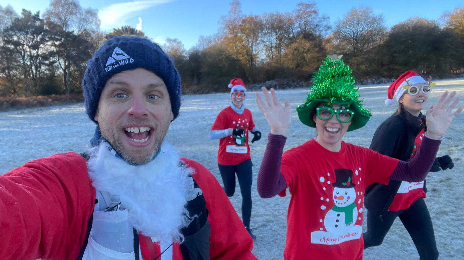 A group of four people dressed in festive holiday gear, including Santa hats, a Santa beard, a Christmas tree hat, and novelty glasses, are running outdoors on a frosty morning. They are smiling and waving at the camera, with trees and a clear sky in the background.