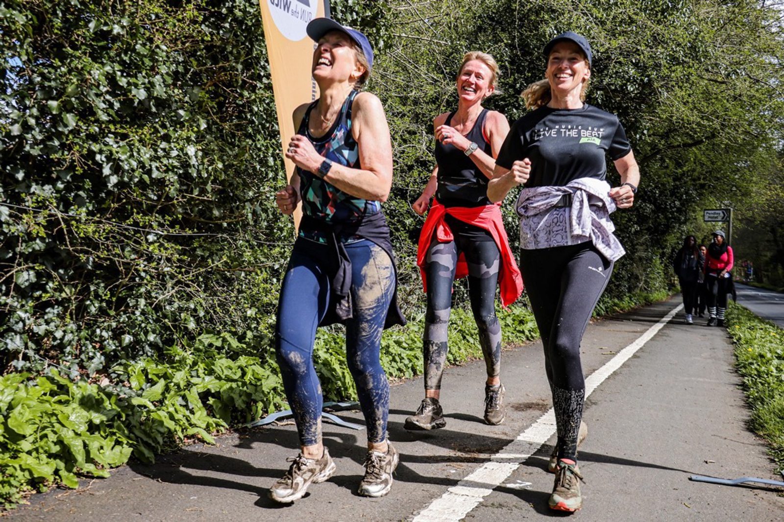 Three women are running outdoors on a sunny day, smiling and covered in mud. They are dressed in athletic clothing, with two wearing caps. They appear to be on a green, leafy path, participating in a race or fitness event. Other people can be seen in the background.