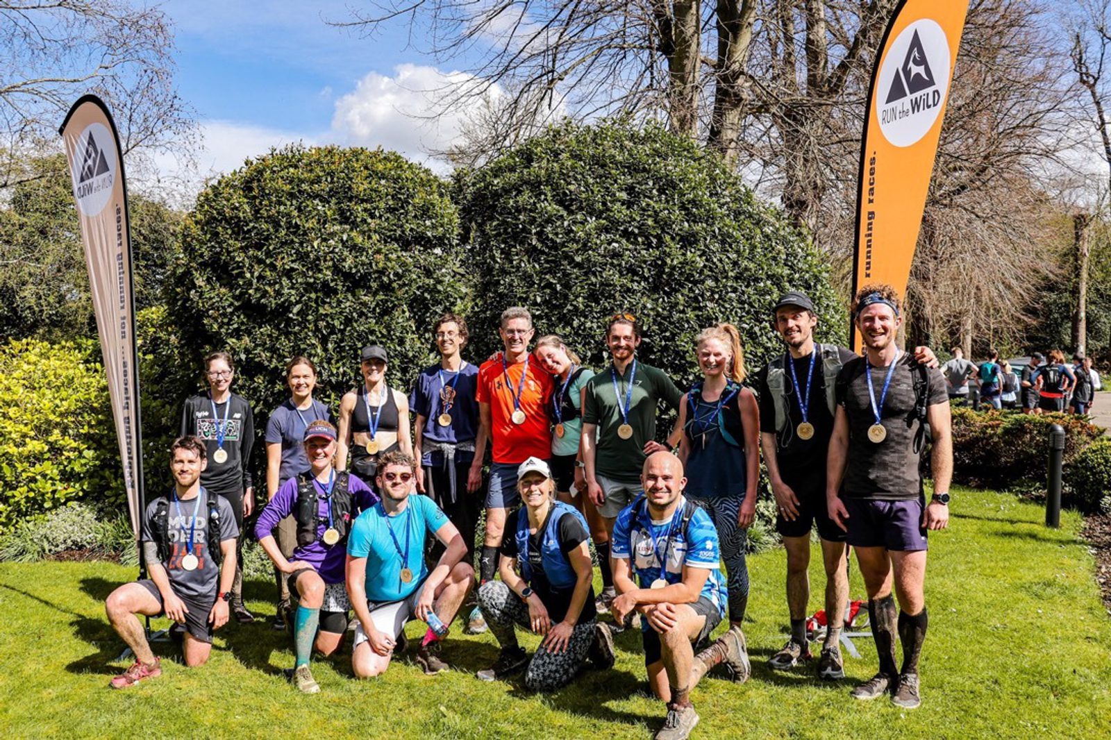 A group of 15 people posing outdoors in front of bushes, some holding medals and wearing race bibs. Two tall banners with the word "Run Wild" and an animal paw logo are on either side. They appear to have participated in a race or outdoor event.