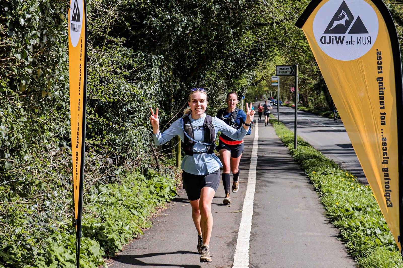 Two runners smile and gesture with peace signs while running on a road flanked by greenery, during a "Run the Wild" event. Yellow event flags and a few other runners are visible in the background.