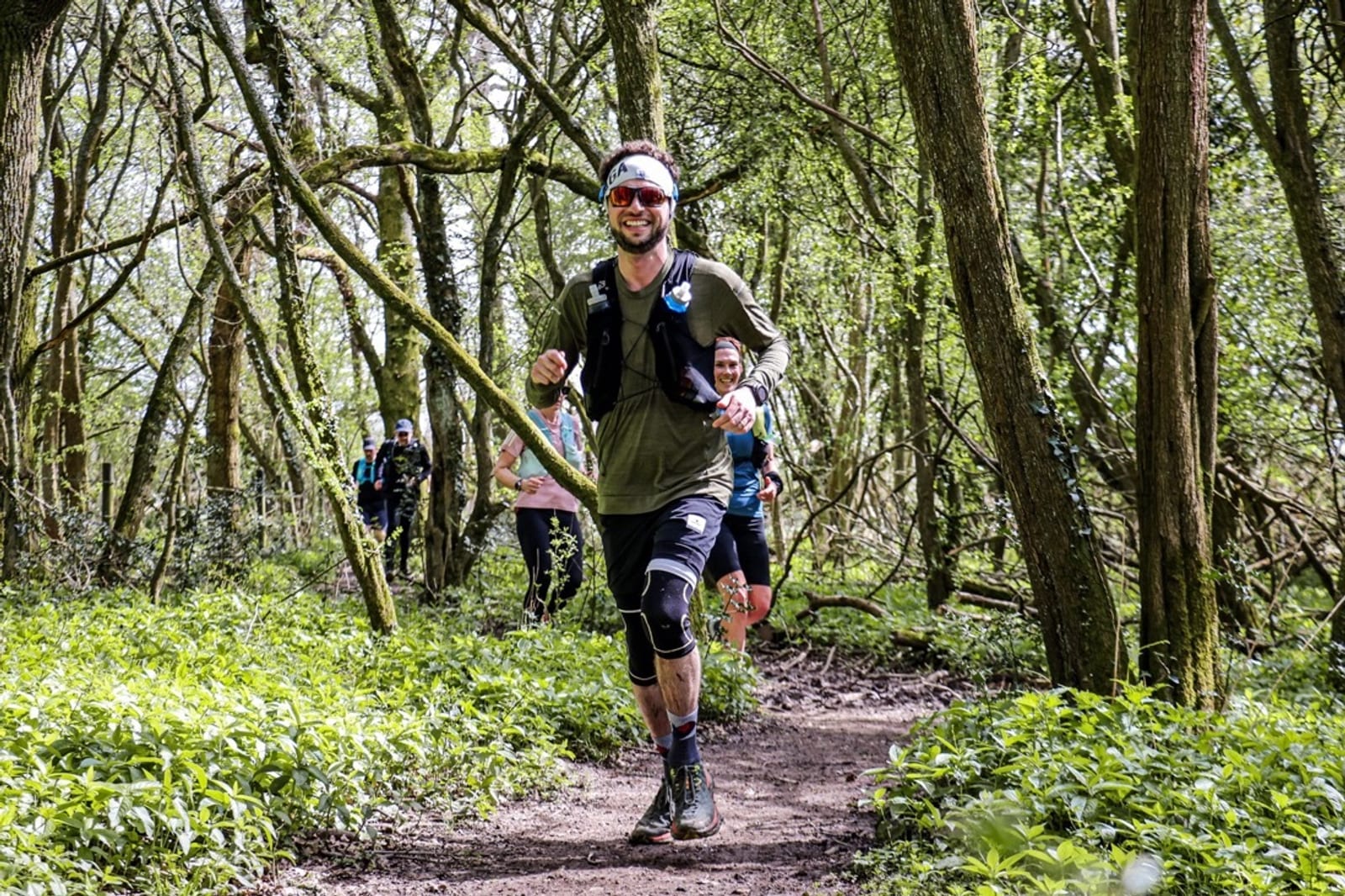 A man wearing sunglasses, a headband, and knee supports runs on a forest trail, smiling, with several other runners following behind him among tall trees and green foliage.