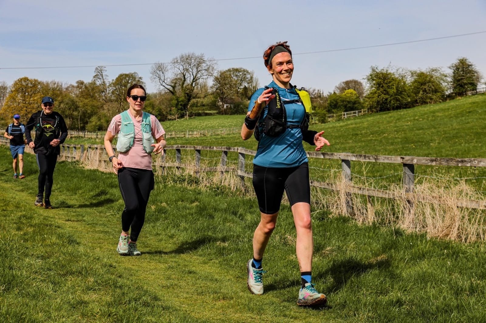 Three people run along a grassy path next to a wooden fence in a rural area. The woman in front smiles, while two others follow behind. Trees and fields are visible in the background under a clear sky.