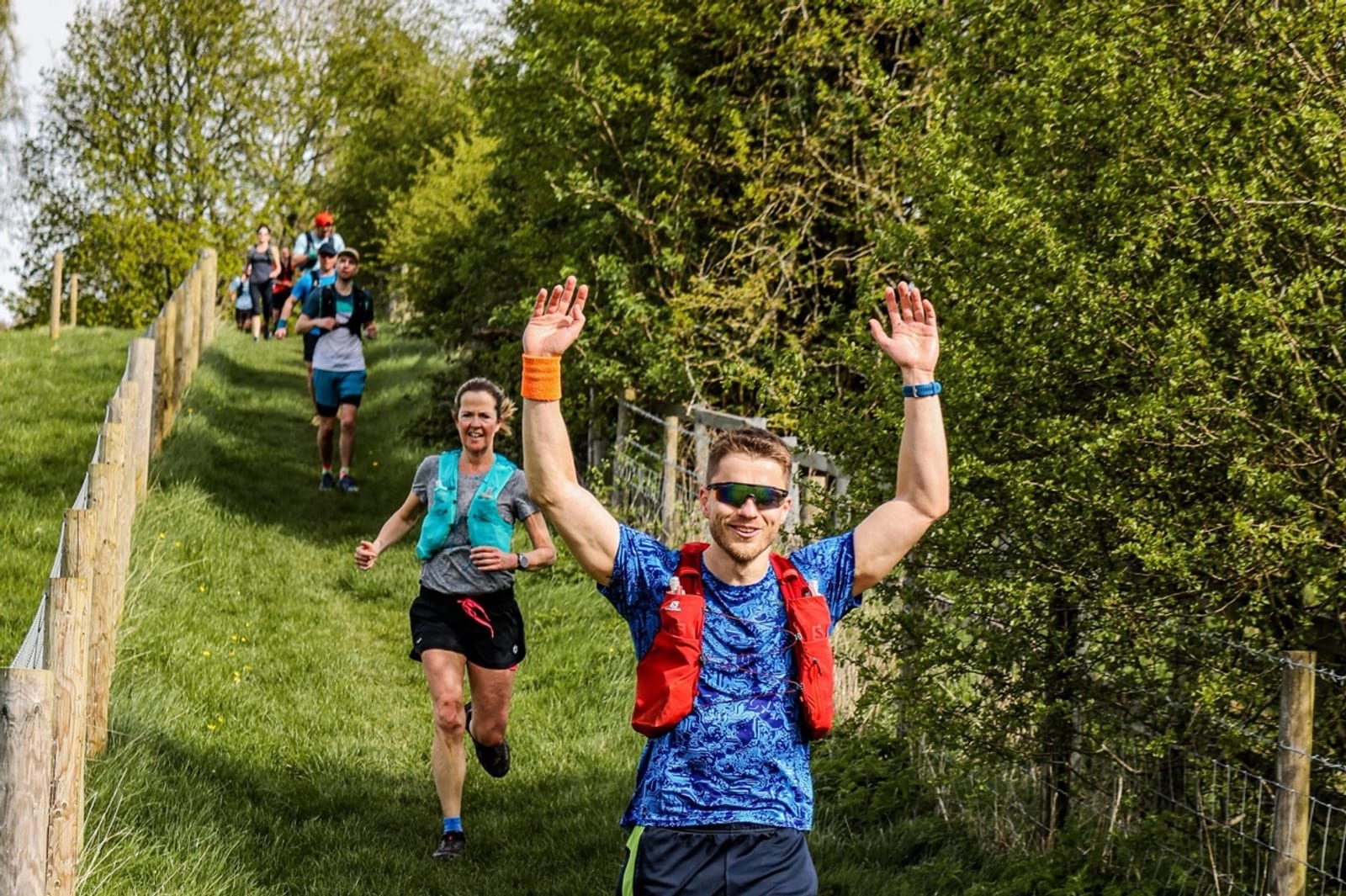 A group of runners in athletic gear jogs down a grassy path beside a fence and trees. The man in front, wearing sunglasses and a red vest, raises his arms and smiles at the camera.