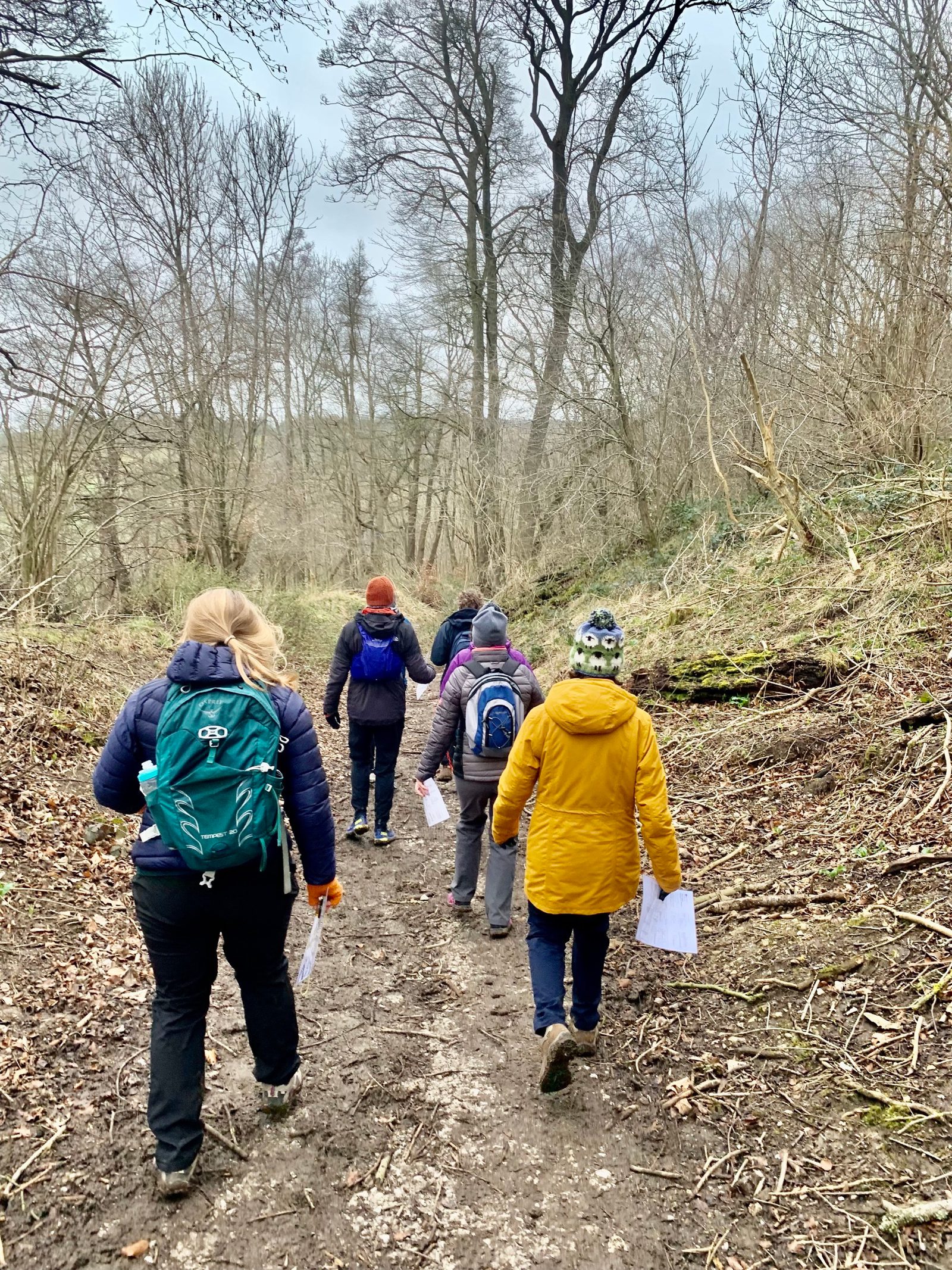 Five people, dressed in winter clothing with backpacks and holding maps, walk down a muddy, tree-lined trail through a forest. The sky is overcast, and there are no leaves on the trees, suggesting a late autumn or winter setting.