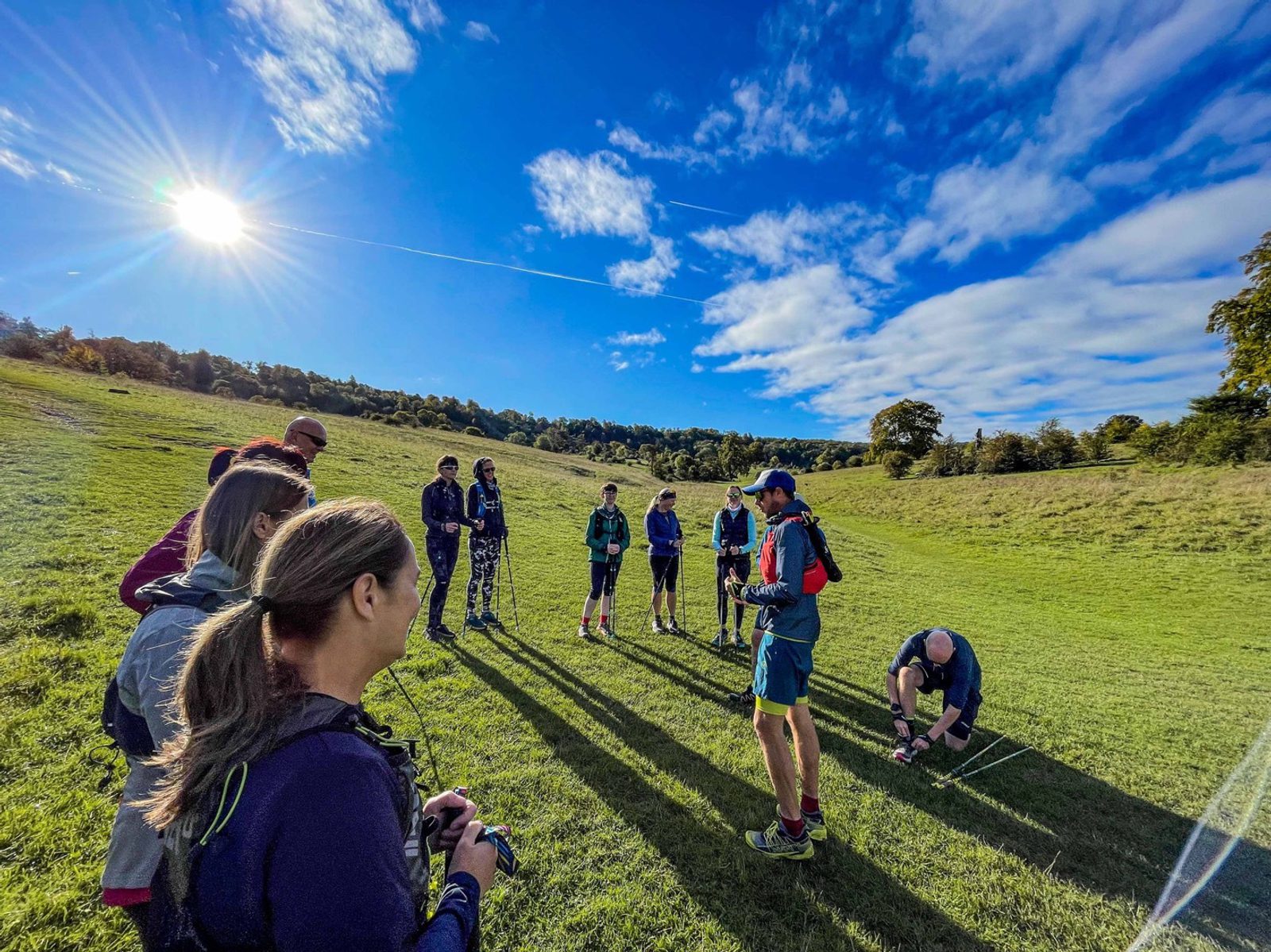 A group of people stands in a sunny field with clear skies, participating in an outdoor activity. One person is writing or drawing on the ground with chalk while others look on, some holding devices or gear, with hills and trees in the background.