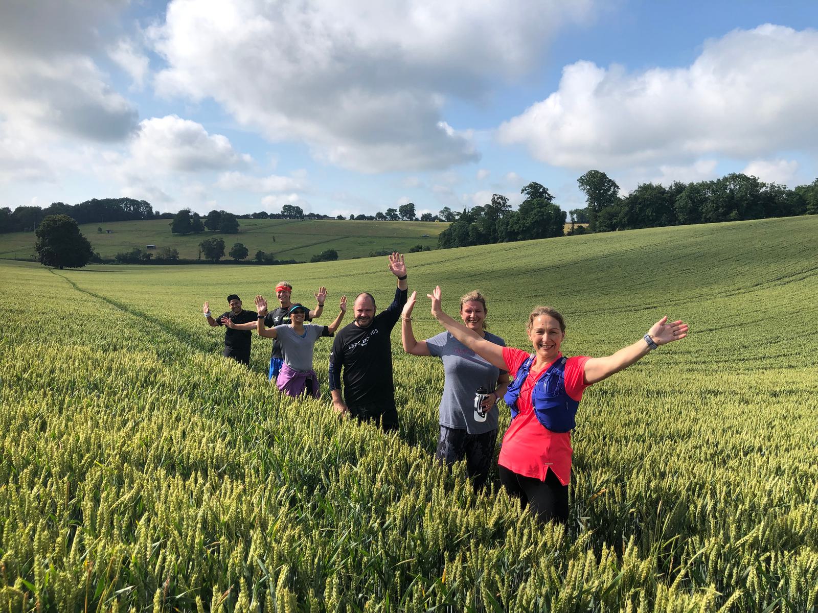 Six people are walking through a lush green field with their hands raised and smiling. The field is surrounded by rolling hills and scattered trees under a partly cloudy sky. They appear to be enjoying a hike or outdoor adventure.