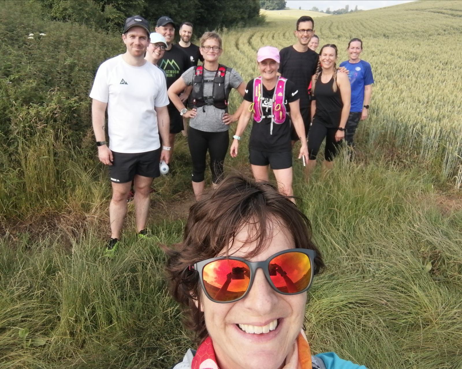 A group of nine people, dressed in athletic attire, pose together on a grassy trail in a rural outdoor setting. One person in the front, wearing sunglasses, takes a selfie while the others smile and stand behind. Fields and greenery stretch out in the background.
