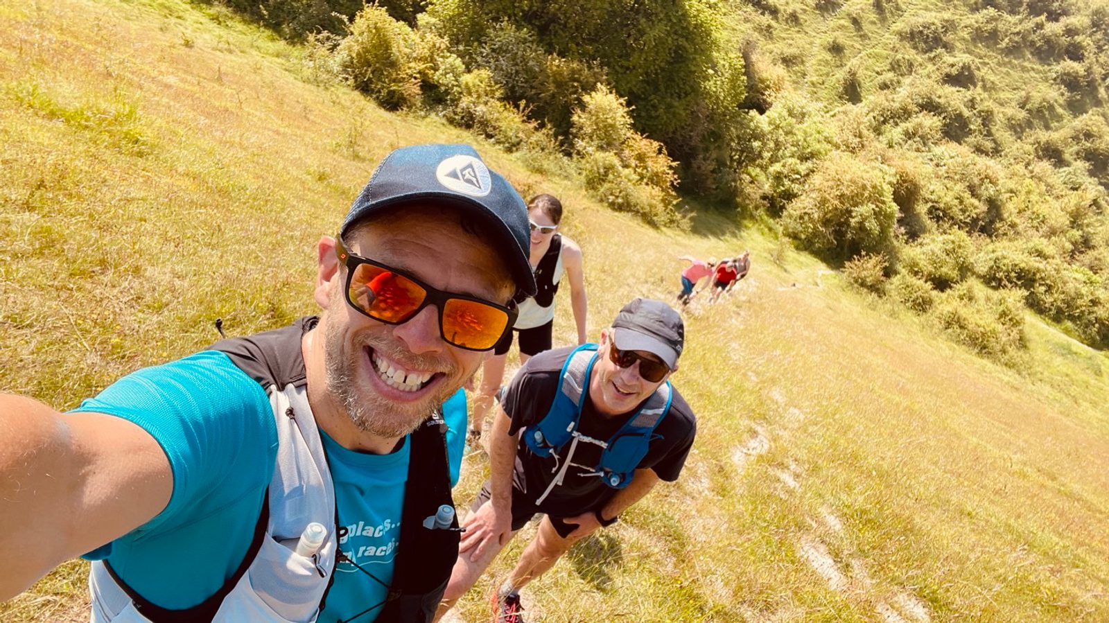 A group of people are hiking up a grassy hillside on a sunny day. The person in the foreground is smiling, wearing a blue cap, sunglasses, and a hydration pack over a blue shirt. The others are further back along the trail, some with similar hiking gear. Trees line the hill.
