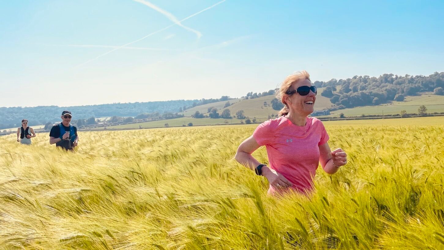 A woman in a pink shirt is running through a field of tall grass on a sunny day. She is smiling and wearing sunglasses and a smartwatch. Three other runners are in the background, with green hills and a clear blue sky visible.