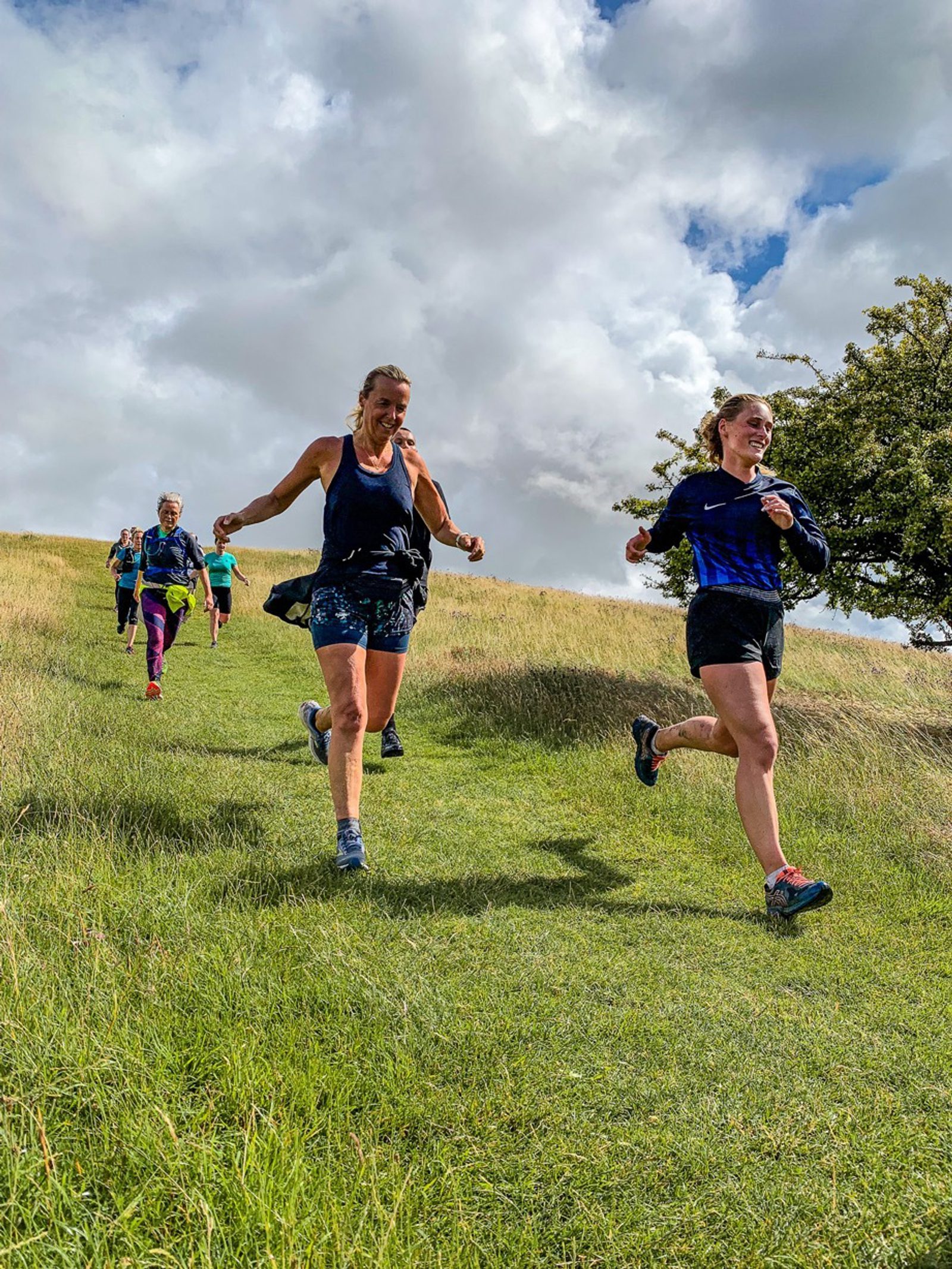 A group of people are running down a grassy hill on a sunny day with some clouds in the sky. The individuals look cheerful and are wearing athletic clothing. In the foreground, two women are smiling and appear to be enjoying themselves. Trees and more runners are visible in the background.
