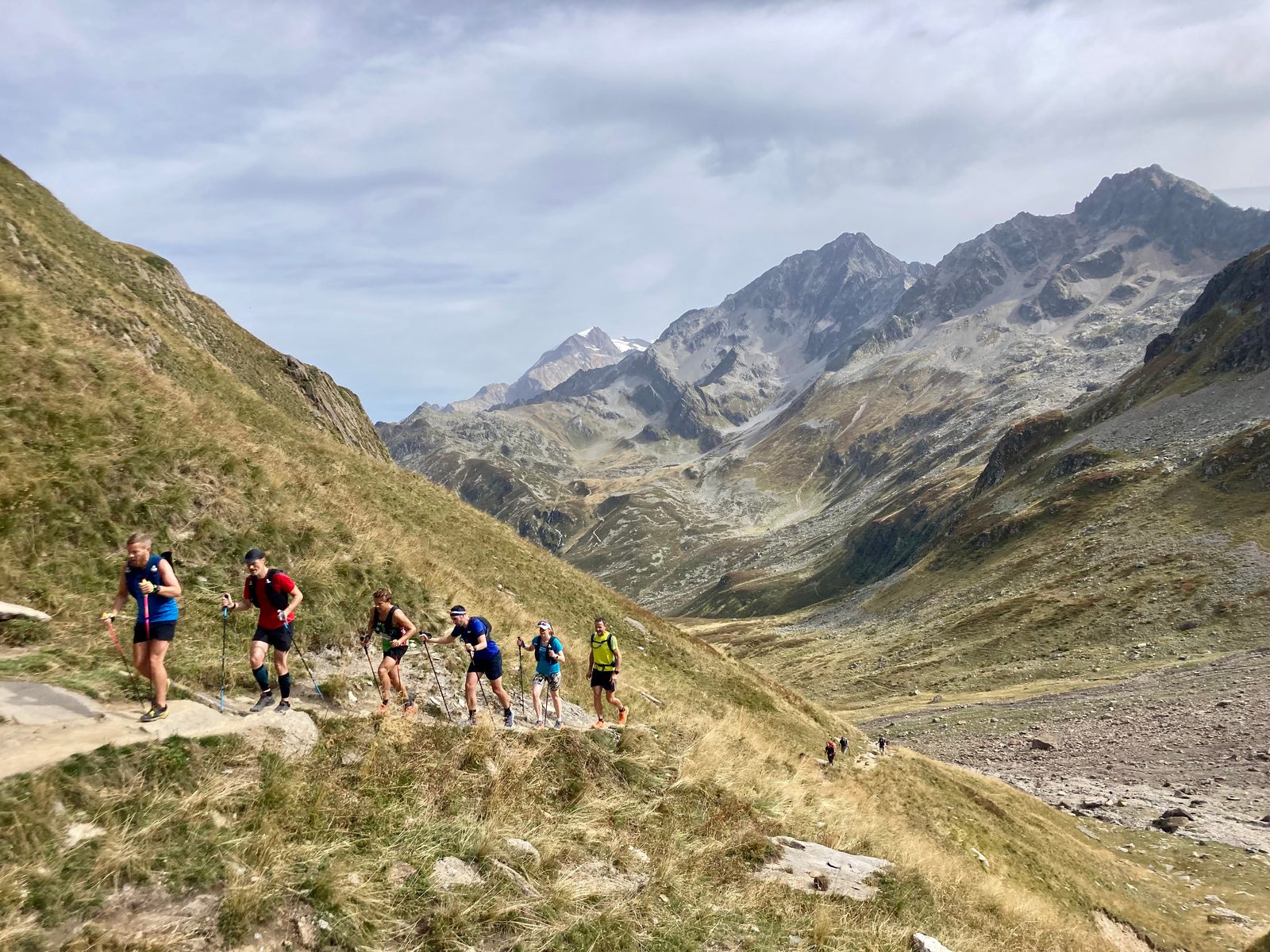 A group of hikers is walking along a trail on a grassy, mountainous landscape. The sky is partly cloudy, and towering rocky peaks are visible in the background. Some hikers are wearing backpacks, and the terrain is uneven, with hills and valleys.