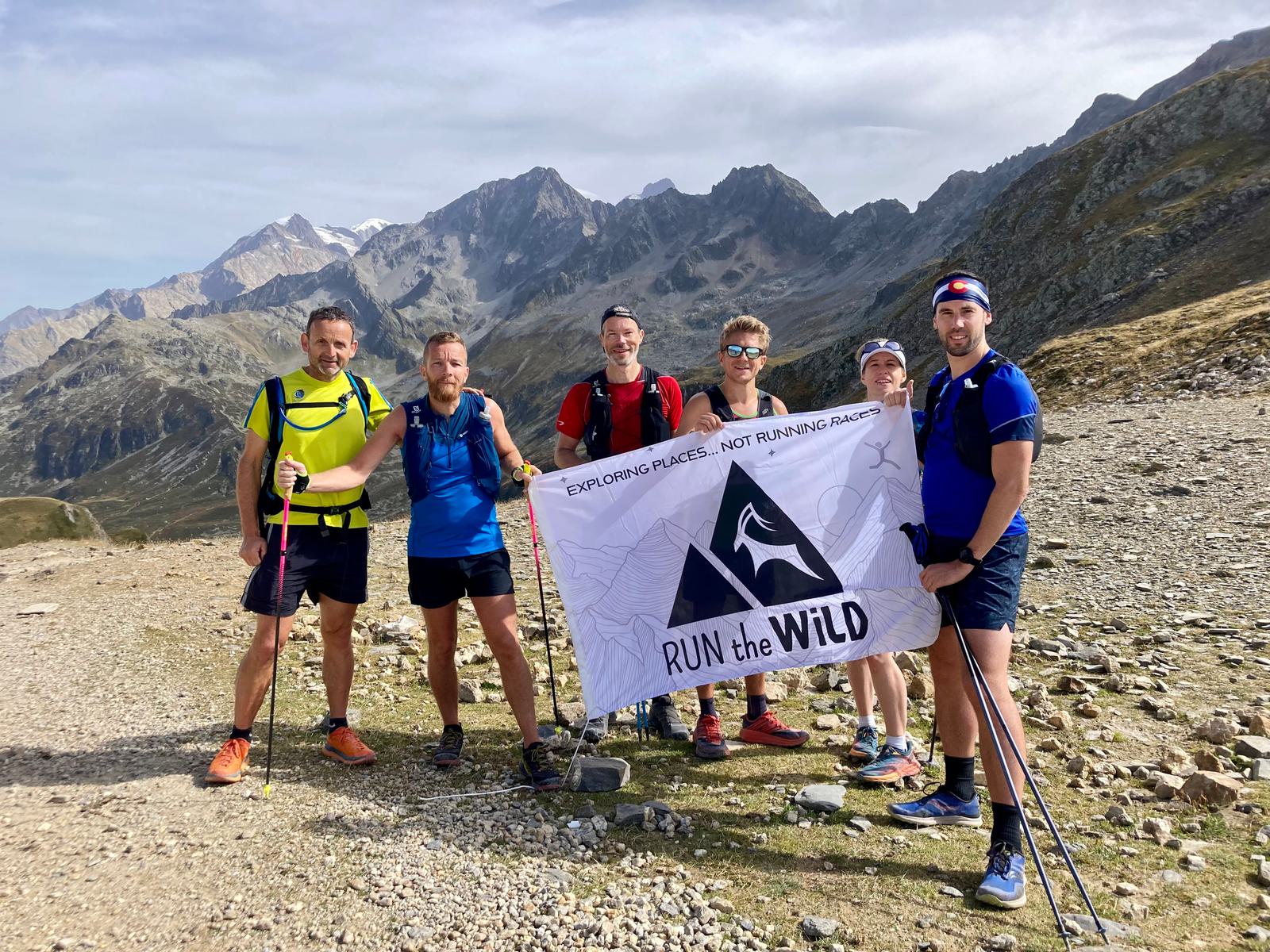 A group of six people in athletic gear stand on a rocky path in a mountainous landscape. They hold a banner that reads "RUN the WILD – EXPLORING PLACES... NOT RUNNING RACES" with a mountain logo. The group smiles at the camera with treks in hand, surrounded by rugged peaks.