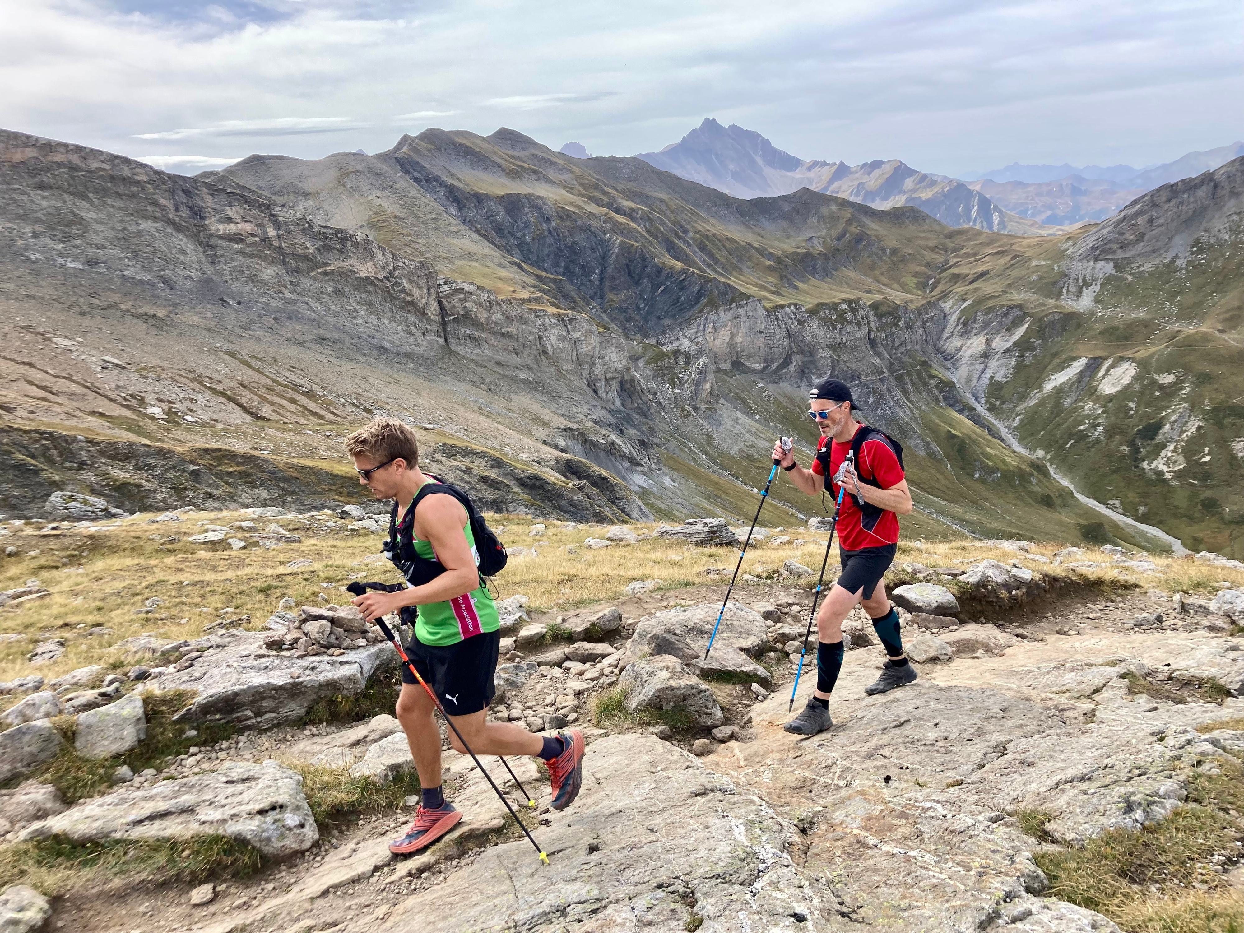 Two trail runners ascend a rocky mountain path with trekking poles. The runner in front wears a green and black outfit, while the one behind sports a red and black outfit with a headband. Majestic mountains and valleys stretch out in the background under a partly cloudy sky.