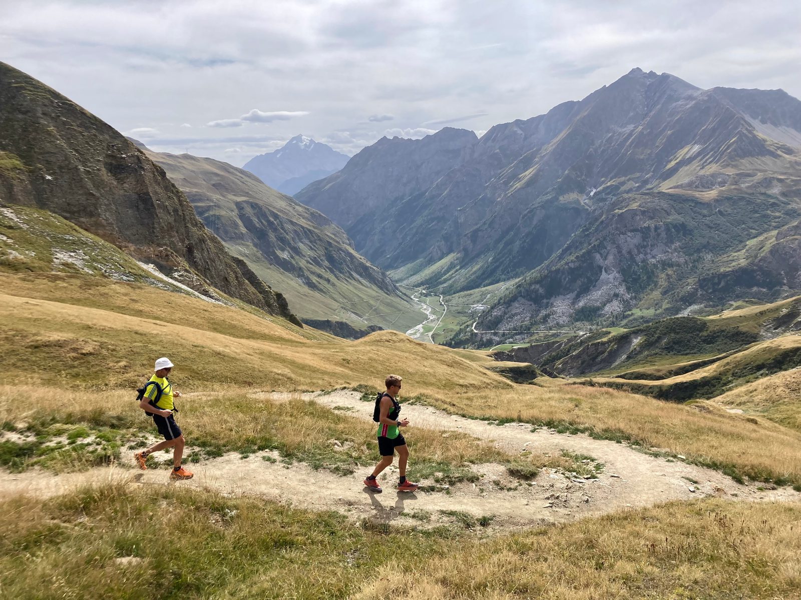Two runners traverse a winding trail through a mountainous landscape with green and brown grassy slopes. Majestic peaks rise in the background under a cloudy sky, and a river can be seen flowing through a distant valley.