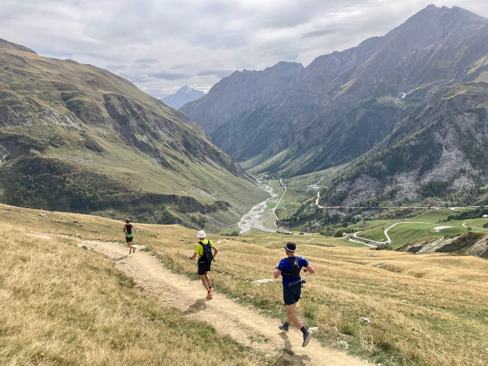 Three trail runners are descending a mountain path surrounded by vast valleys and towering peaks. The sky is overcast, and the lush green landscape below contrasts with the rocky terrain. A narrow river winds through the valley.