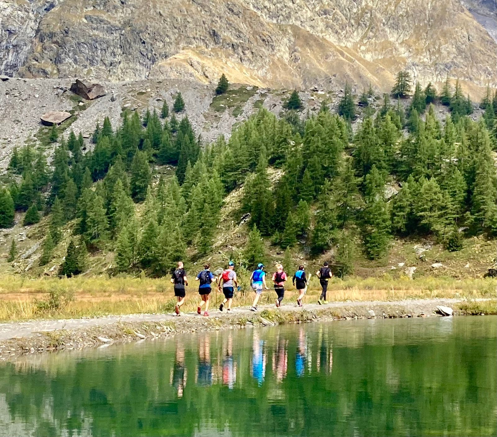 A group of six people wearing hiking gear walks along a narrow path beside a reflective lake in a mountainous area. The background shows rugged mountains with greenery, featuring a mix of trees and rocky terrain. The scene is bright and scenic.