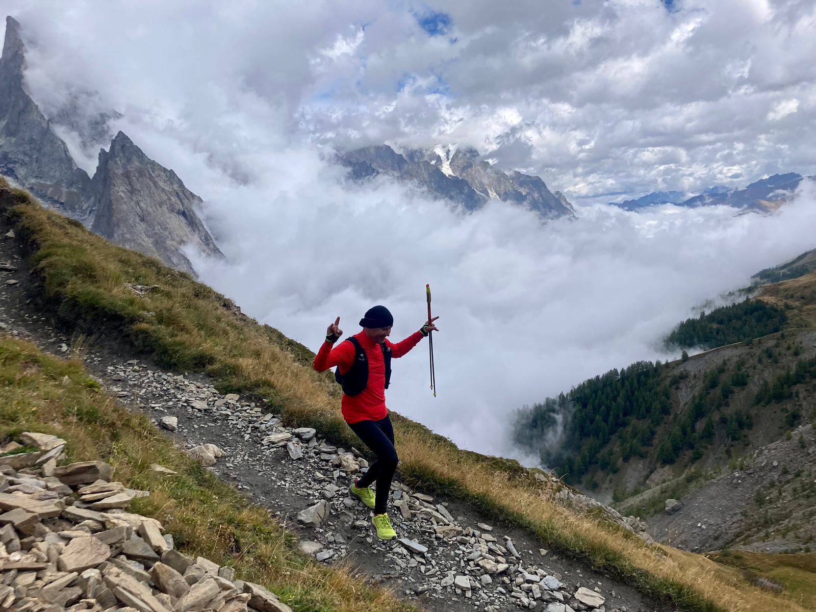 A person wearing a red jacket, black pants, and yellow shoes is running on a rocky, mountainous trail with trekking poles in both hands. They are surrounded by cloudy skies and distant mountain peaks. The trail overlooks a lush valley covered in greenery and clouds.