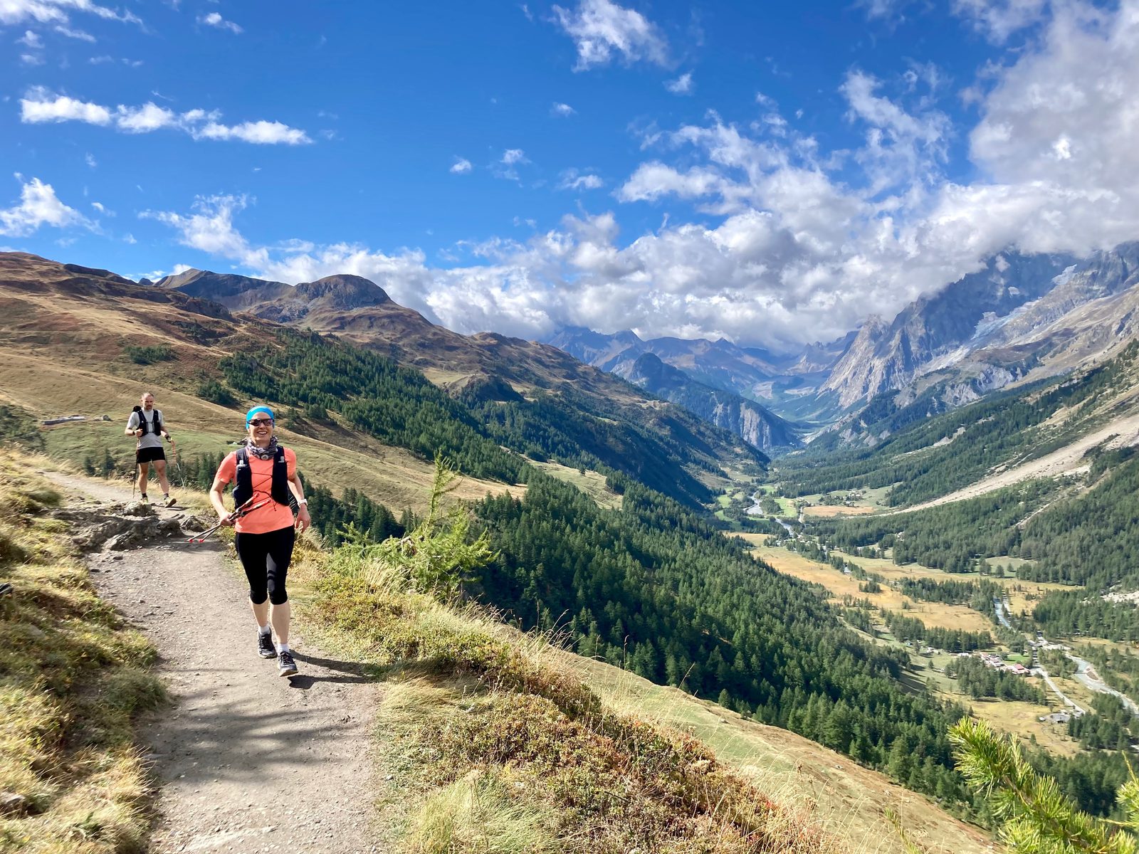 Two people run on a dirt trail through a mountainous landscape. The foreground runner wears a pink shirt, black shorts, and a blue hat. Green valleys, rolling hills, and towering peaks fill the background under a partly cloudy sky. Lush forests stretch below.