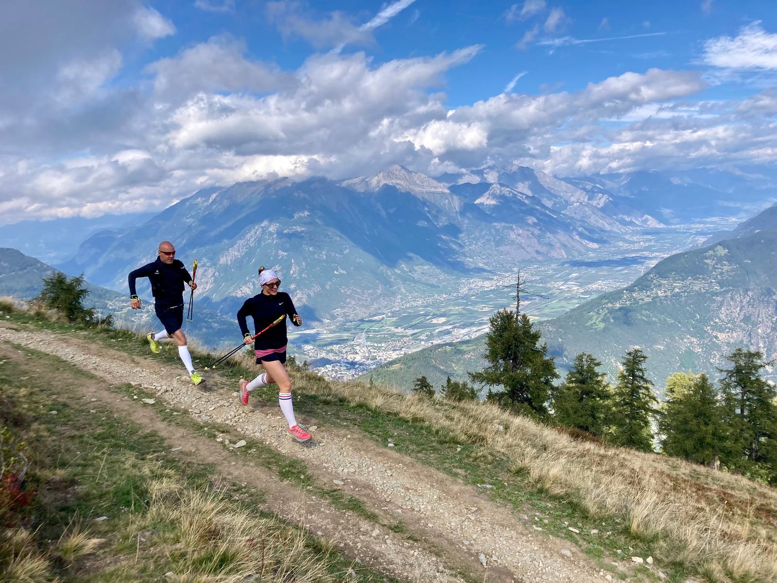 Two trail runners, one in blue and the other in black, run along a rugged mountain path with trekking poles. They are surrounded by green hills and tall trees, with a breathtaking view of a vast valley and distant mountain range under a partly cloudy sky.