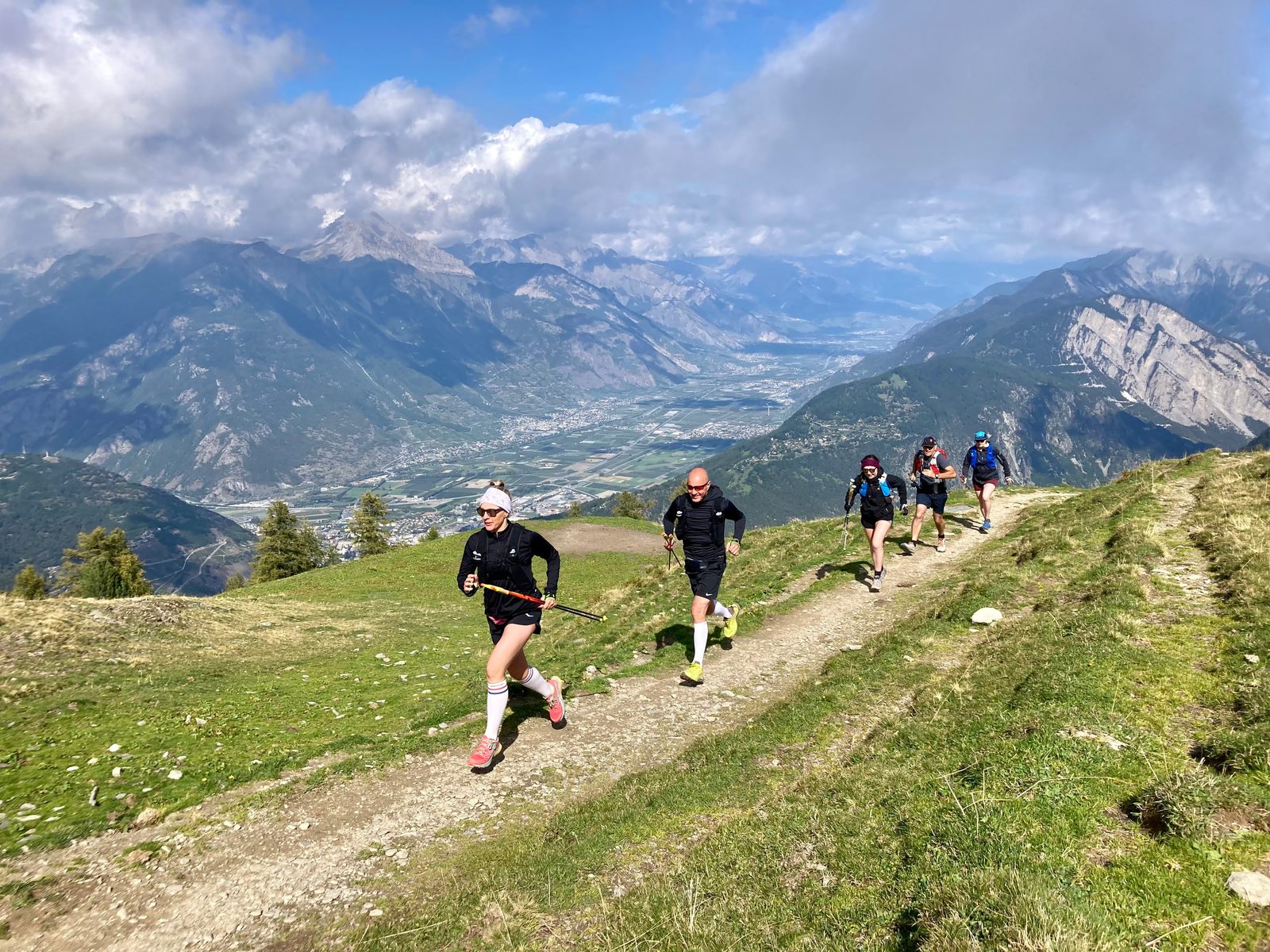 Five people are trail running up a dirt path on a mountainside covered in green grass. The background reveals a vast valley surrounded by towering peaks and partly cloudy skies. The runners are dressed in athletic gear, and the scene is energetic and scenic.