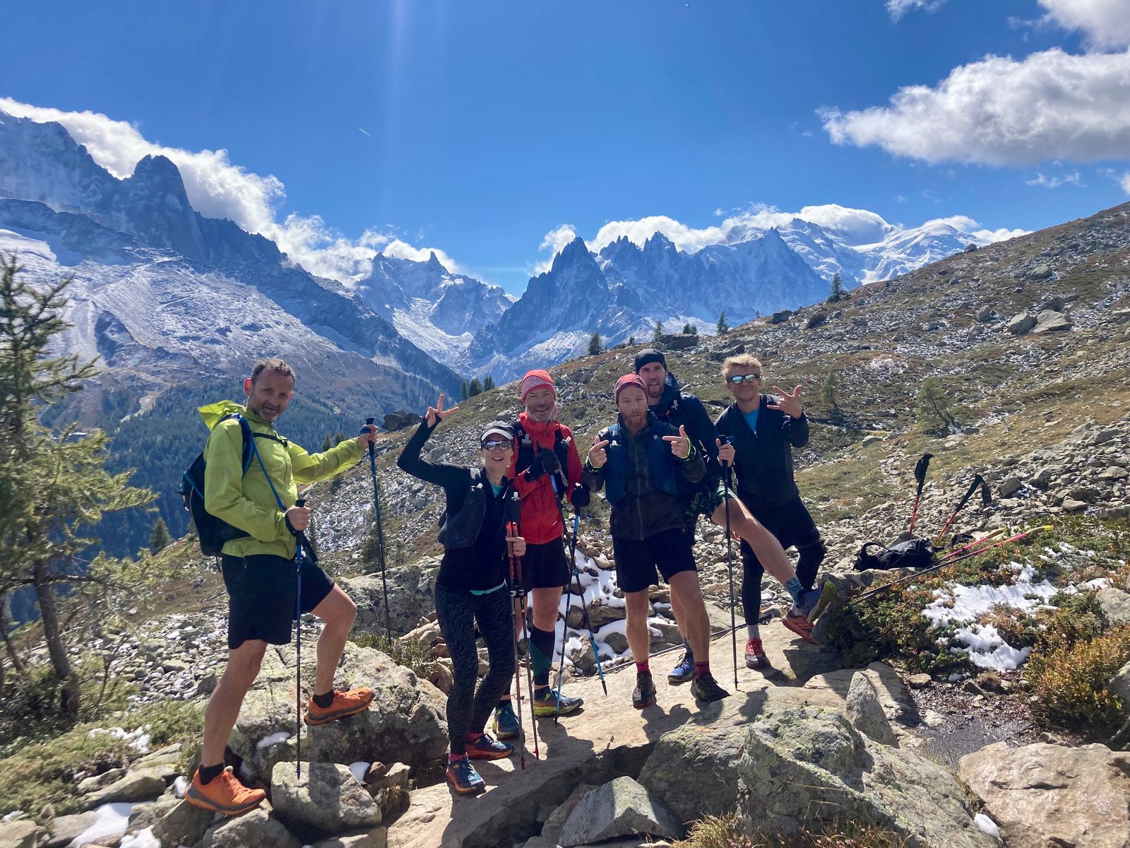 Five hikers, wearing outdoor gear and carrying trekking poles, pose energetically on a rocky mountain trail. Snow-capped peaks and a blue sky with scattered clouds form a stunning backdrop. Vegetation around the hikers indicates a high-altitude environment.