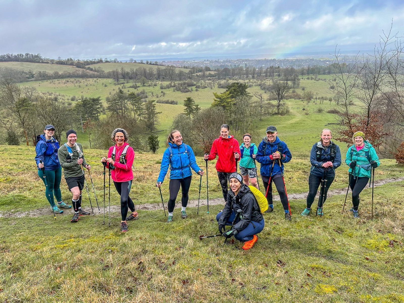 A group of ten people dressed in outdoor gear pose with trekking poles on a grassy hilltop with a scenic backdrop of fields and trees. They appear happy and are likely taking a break from hiking. The sky is cloudy with patches of blue visible.