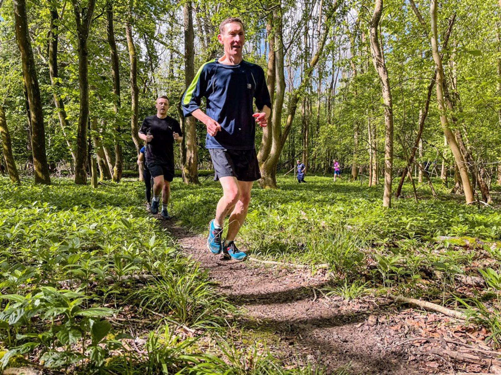Two people are running on a dirt path through a sunlit forest with lush green foliage. The lead runner wears a black and blue outfit, while the second runner has a black outfit. Sunlight filters through the trees, creating a vibrant natural setting.