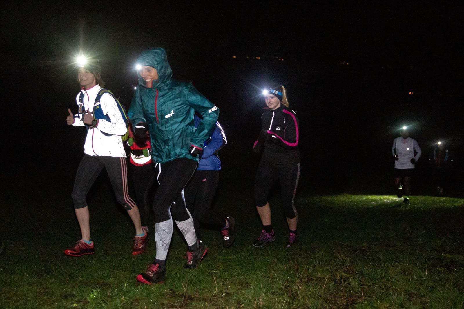 A group of people running outdoors in the dark, wearing headlamps and athletic gear. The grass is visible underfoot, and some distant lights can be seen in the background. The runners are dressed warmly, suggesting cool weather conditions.