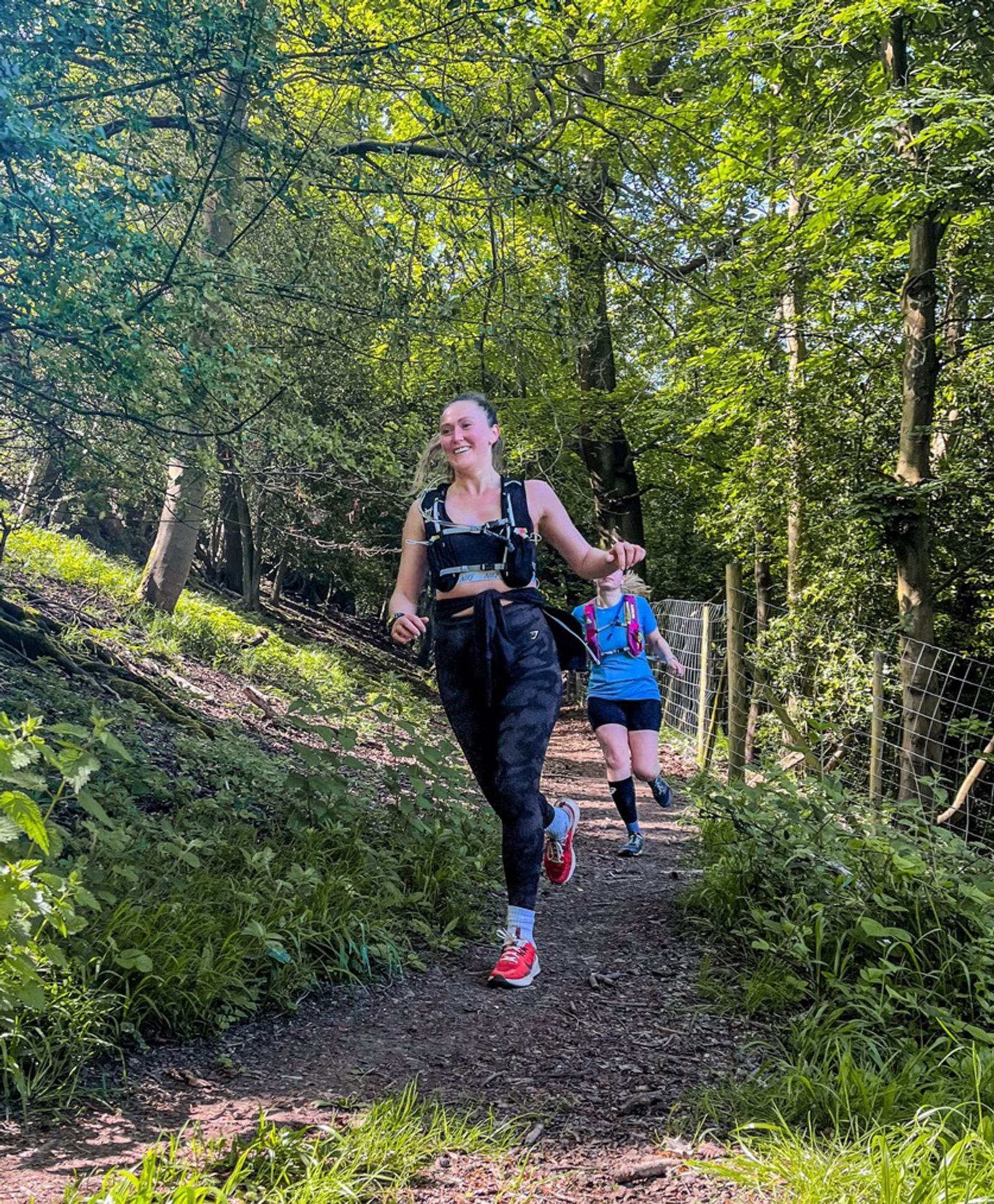 Two people are running along a forest trail. The front runner wears a black outfit and red shoes, and the person behind wears a blue shirt and shorts. The path is surrounded by lush greenery and trees under a sunny sky.