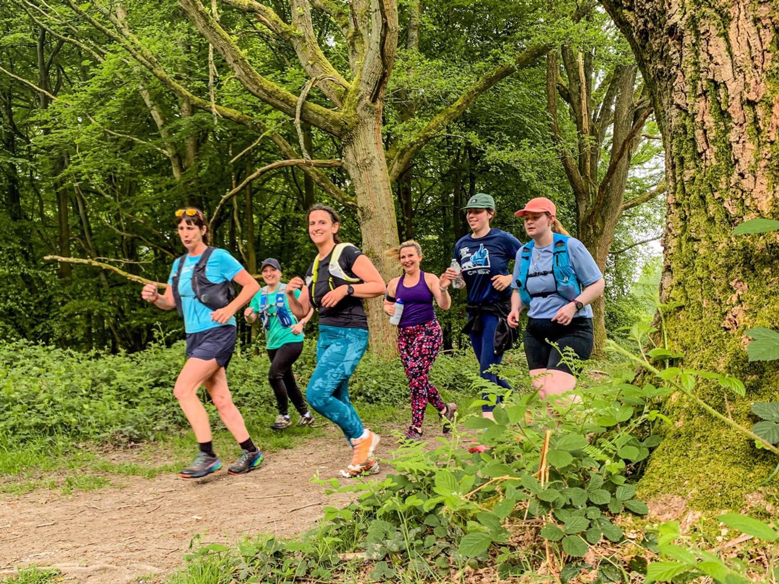 A group of six people is jogging together on a dirt path through a lush, green forest. They are wearing casual athletic clothing and appear cheerful, with trees surrounding them.