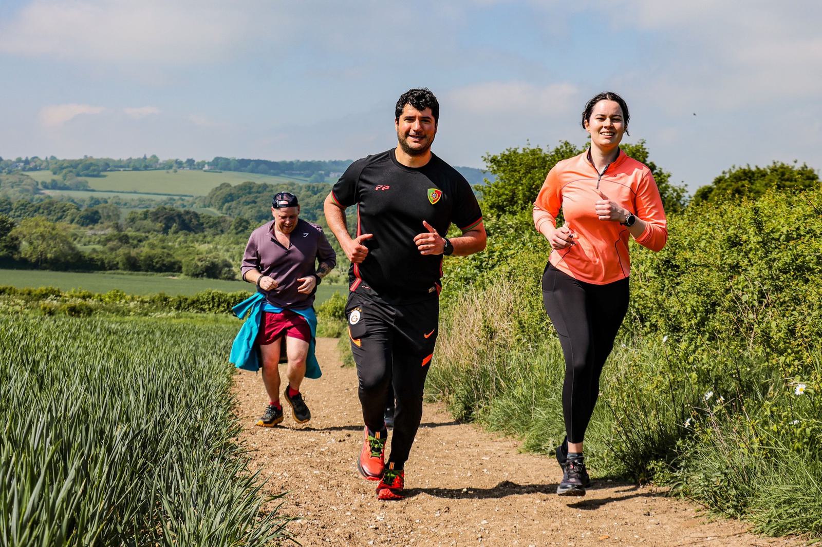 Three people jogging together on a dirt path through a scenic countryside. The runners, two men and a woman, are smiling and appear to be enjoying the sunny day. Lush green fields and blue skies are visible in the background.