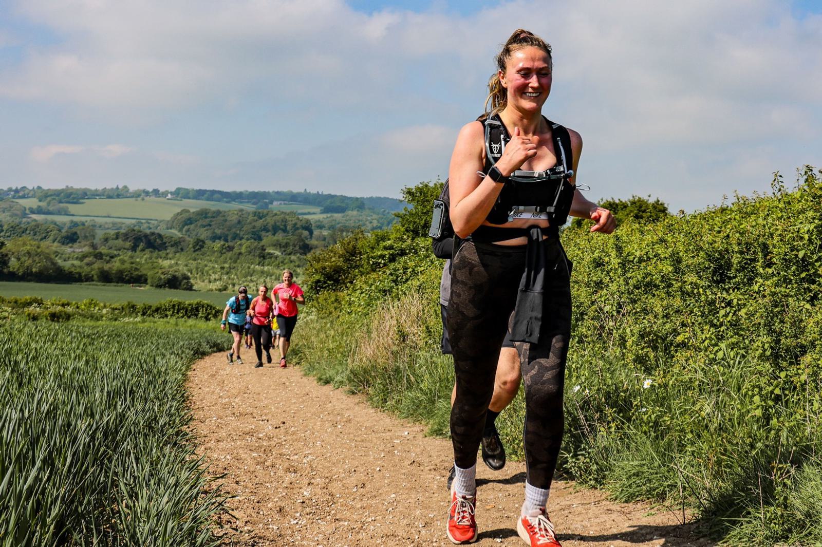 A woman is jogging on a rural dirt path, smiling, wearing athletic gear with a hydration pack. Behind her, a small group of runners follows. The background features lush green fields and hills under a blue sky.
