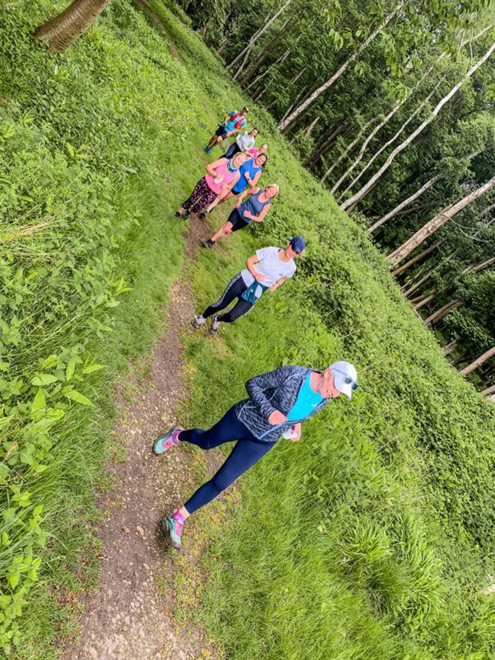 A group of people jogs along a narrow, winding path surrounded by lush green grass and tall trees in a forest. They are wearing athletic clothes and appear to be enjoying the outdoor activity.
