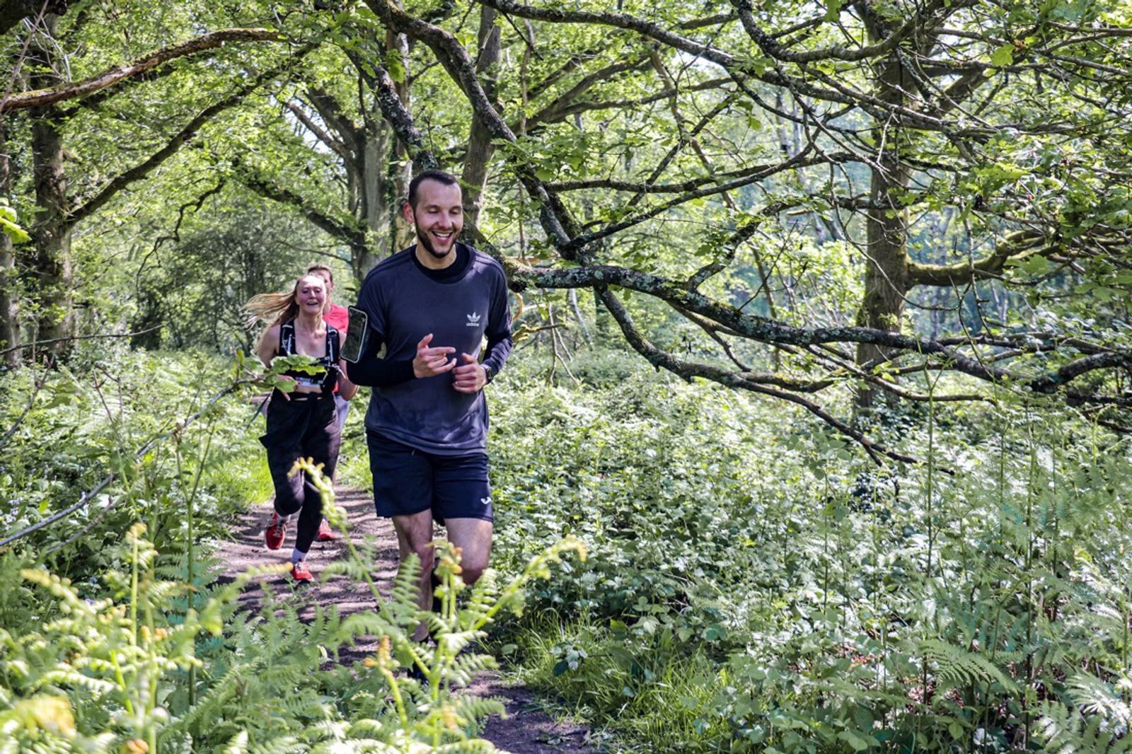 Two people jogging on a narrow, dirt path through a lush, green forest. They both wear black athletic clothing. Sunlight filters through the trees, casting dappled light on the path and surrounding foliage.