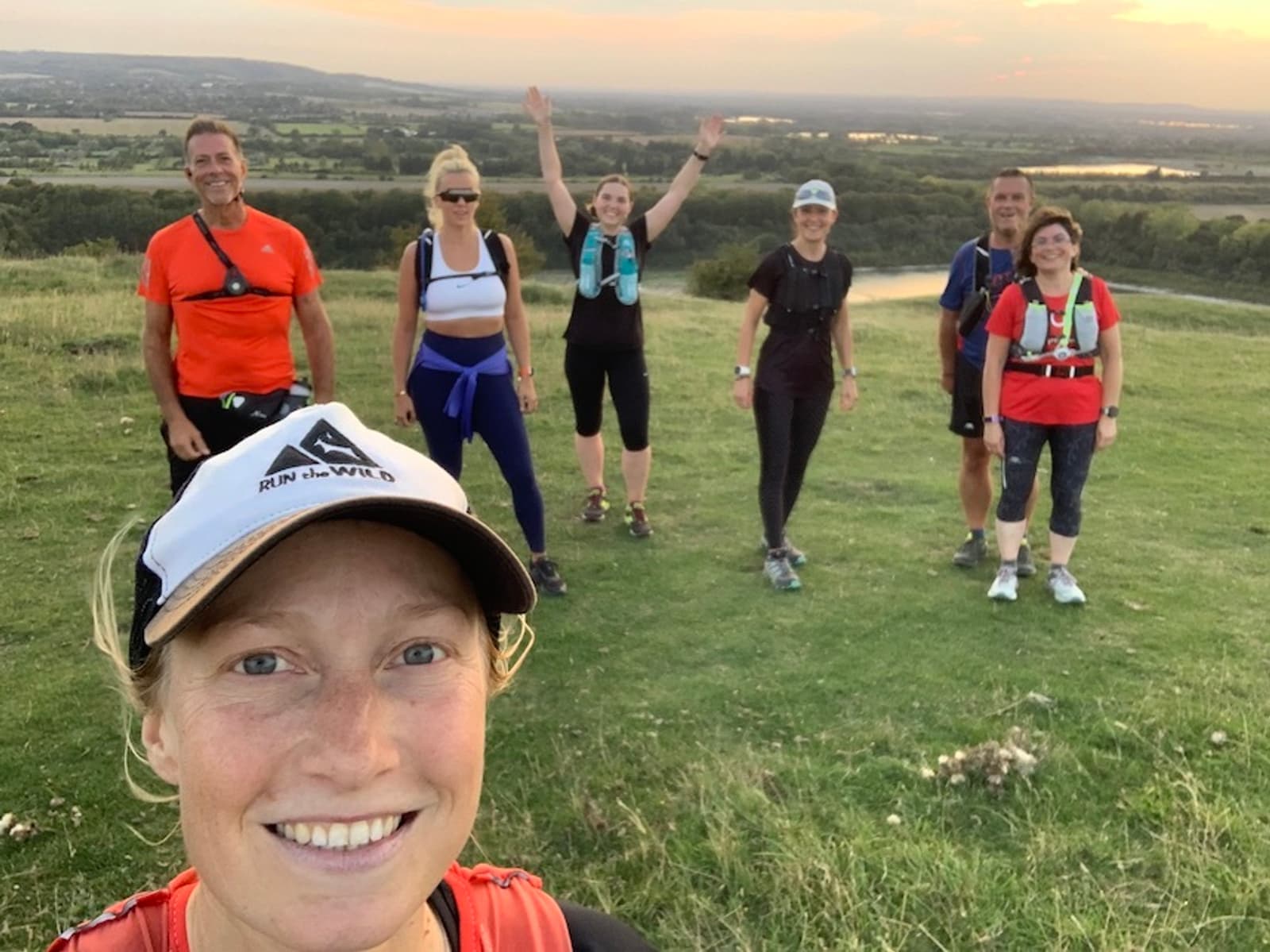 A group of six people standing on a grassy hill, smiling and posing for a photo. The person in front is taking a selfie, wearing a white and black cap. The background shows a scenic landscape with trees and a river, under a colorful evening sky.