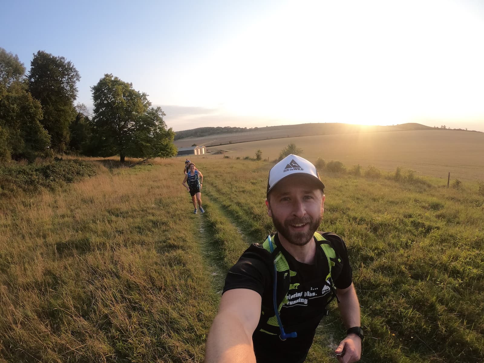 Two people are running on a narrow path through a grassy field at sunset. The person in the foreground is smiling and wearing a black athletic shirt with a hydration pack, while the person behind them is further down the path. Trees and open fields surround them.